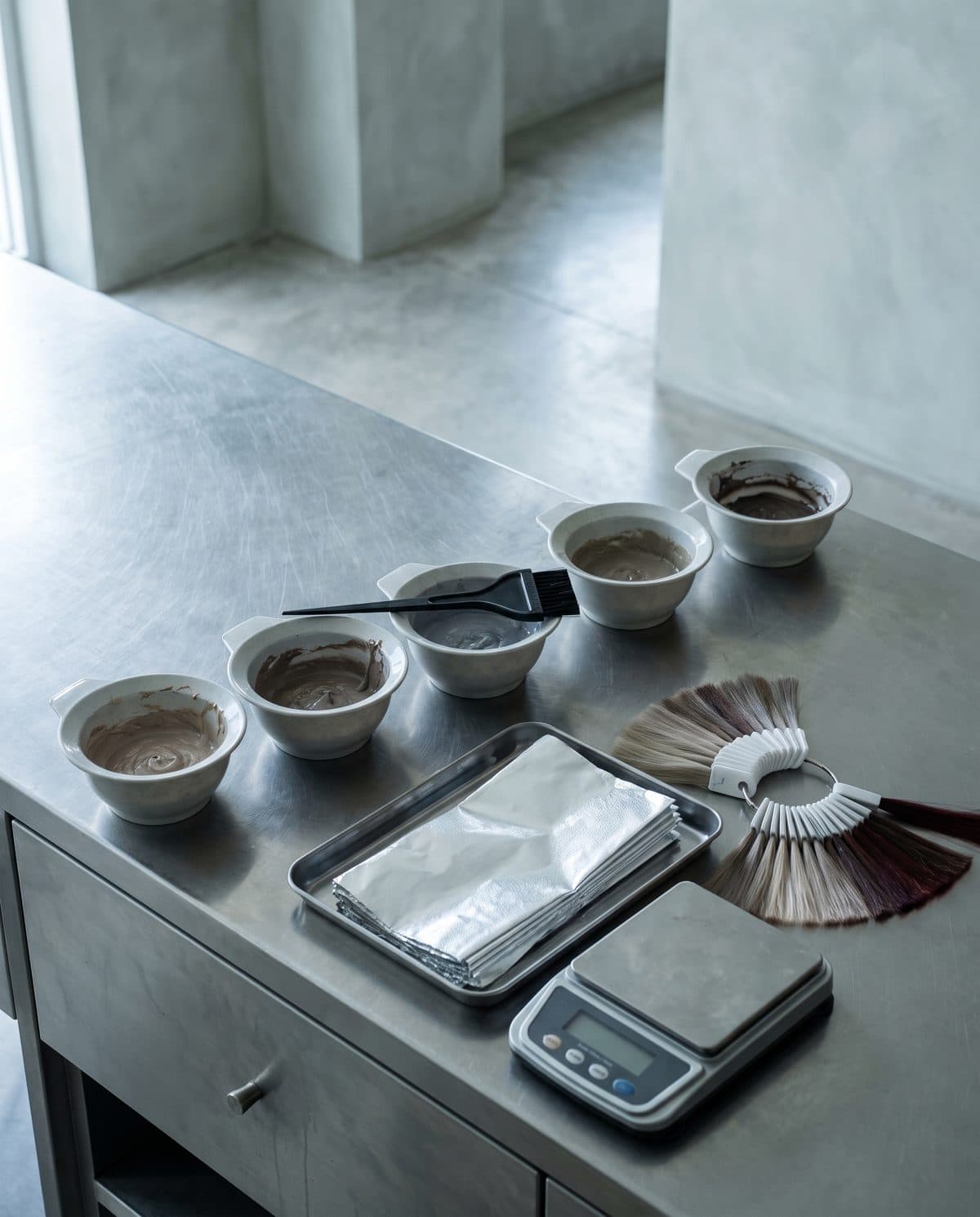 A salon colour bar from above: tint bowls, a brush, folded foils and a swatch ring on a stainless counter